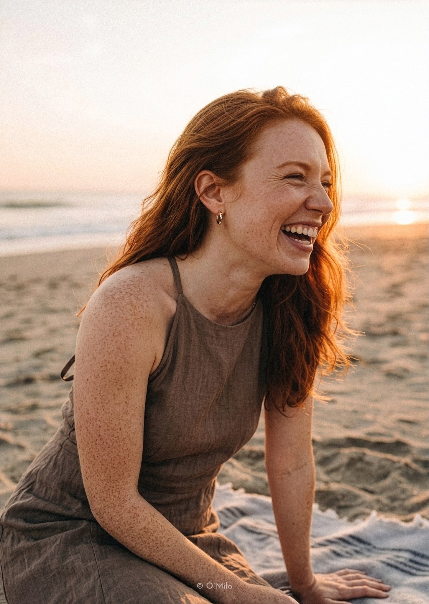 A woman with red hair smiling and laughing on a beach at sunset, wearing a brown sleeveless dress. The background shows sand, ocean, and warm golden light.